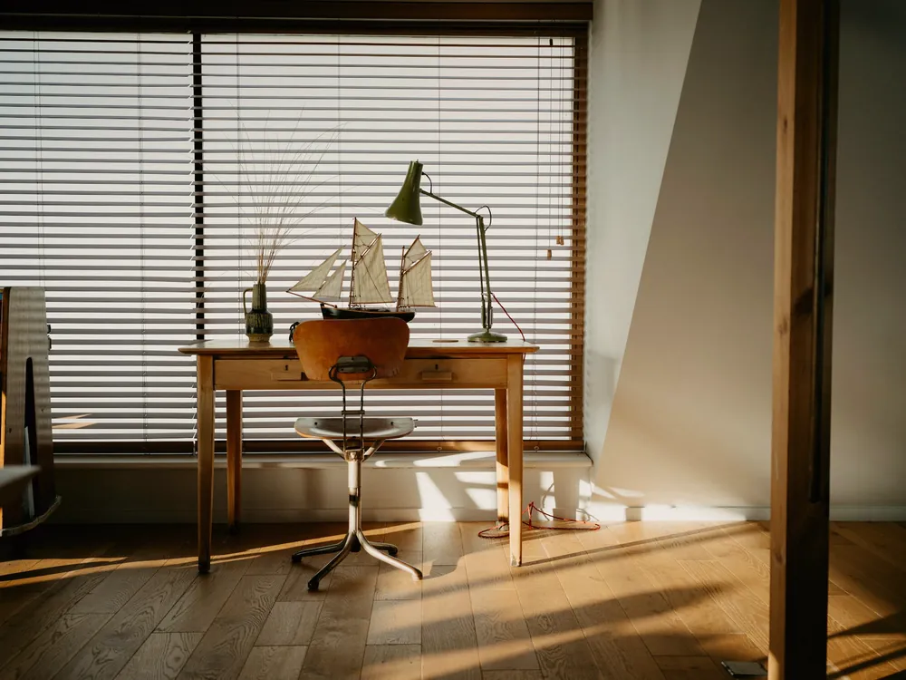 Bureau en bois devant une fenêtre à stores, avec une maquette de voilier, une lampe articulée et un tabouret