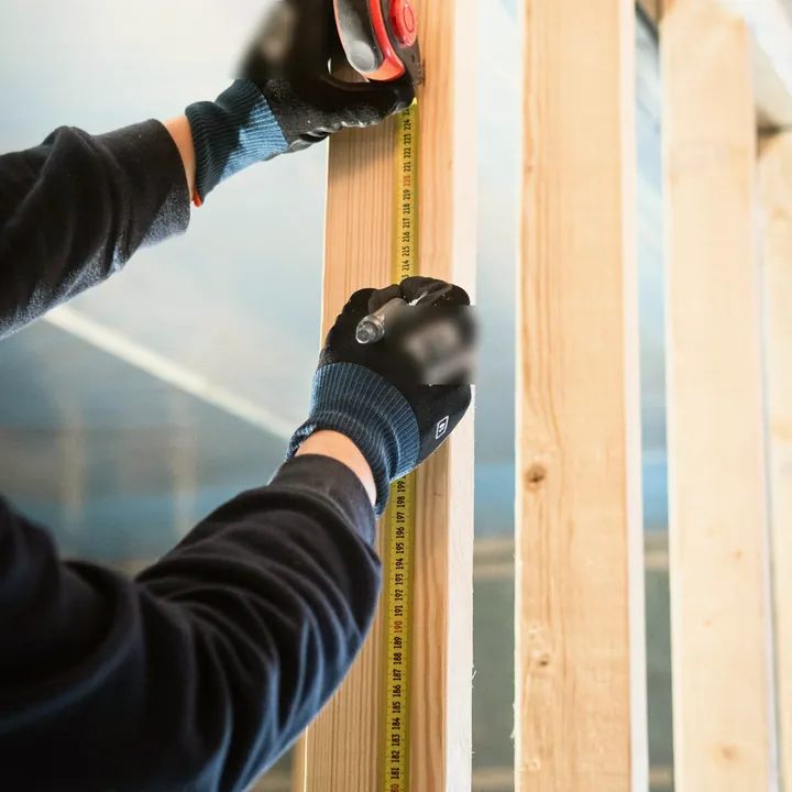Mains gantées déroulant un mètre ruban le long d’un montant en bois, sur un chantier
